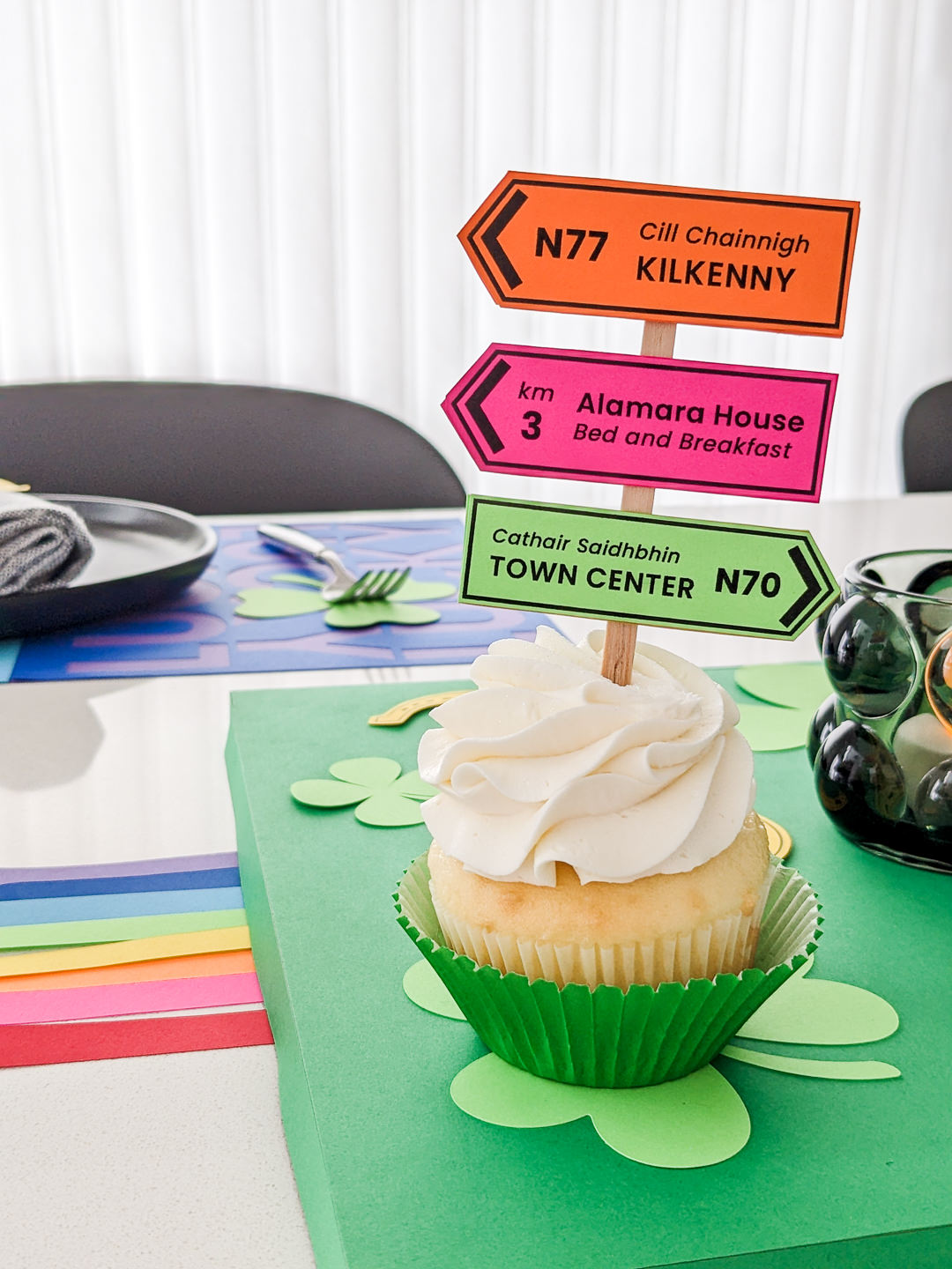 Cupcake with Irish road signs cupcake topper sitting on a shamrock-decorated table for St. Patrick's Day