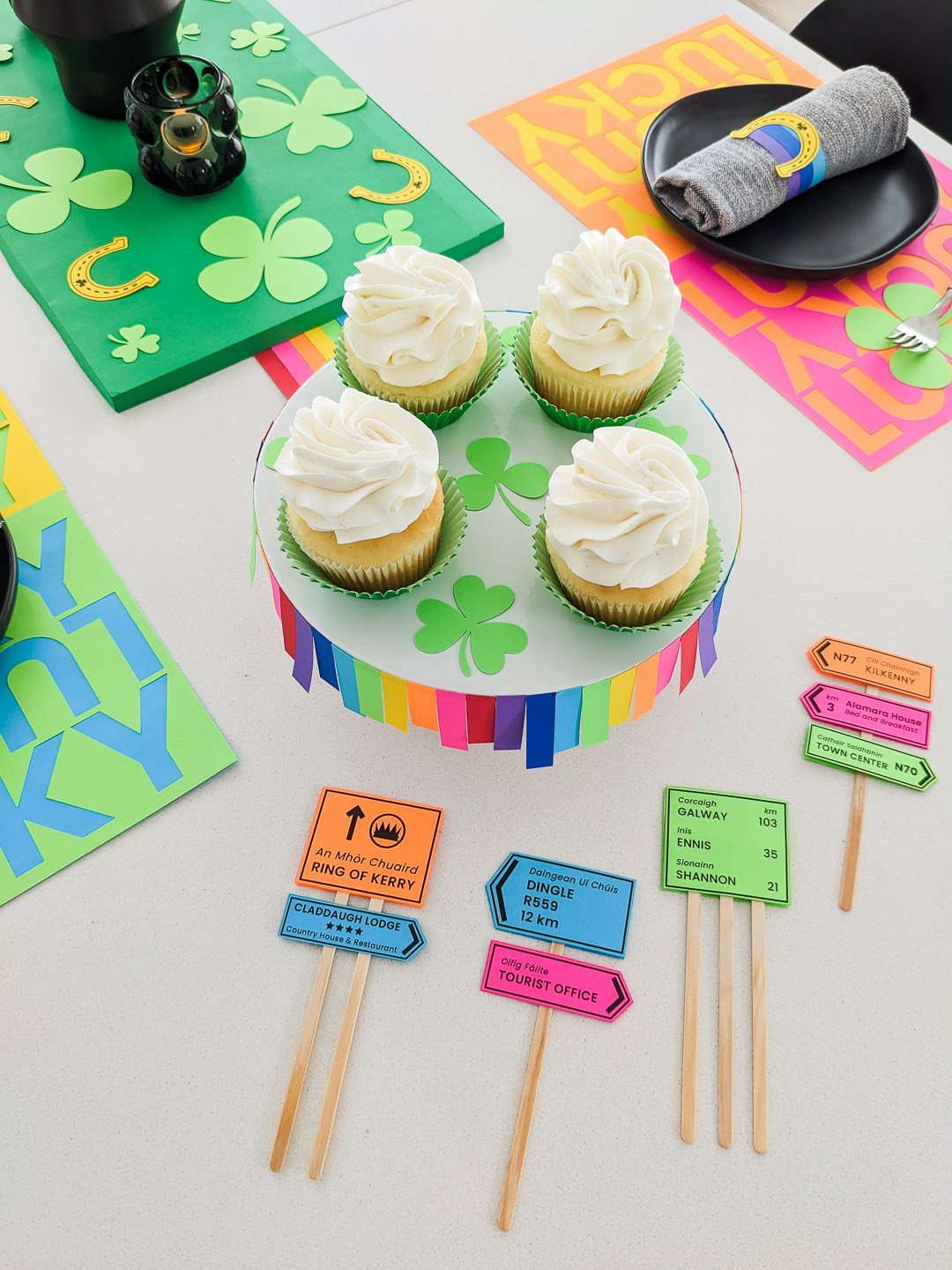 DIY Irish road signs cupcake toppers on a table next to cupcakes sitting on a rainbow cupcake stand with St. Patrick's table decor in the background